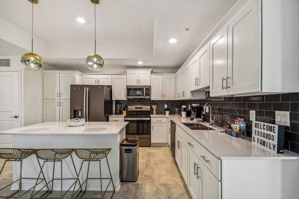 This modern kitchen boasts a spacious island with gleaming white countertops, ideal for gatherings. The rich dark backsplash contrasts beautifully with bright cabinetry, and stylish pendant lights enhance the chic atmosphere.