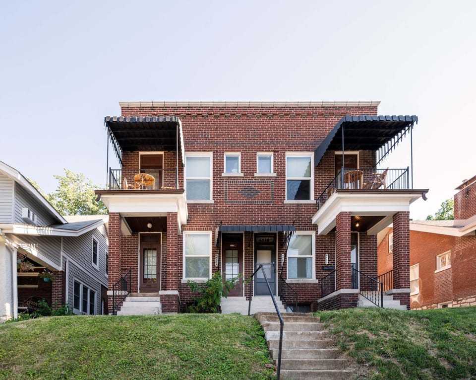 The daytime exterior shows a two-story brick building with multiple front entries, covered porches, upper balconies, central concrete steps, and a sloped front lawn.