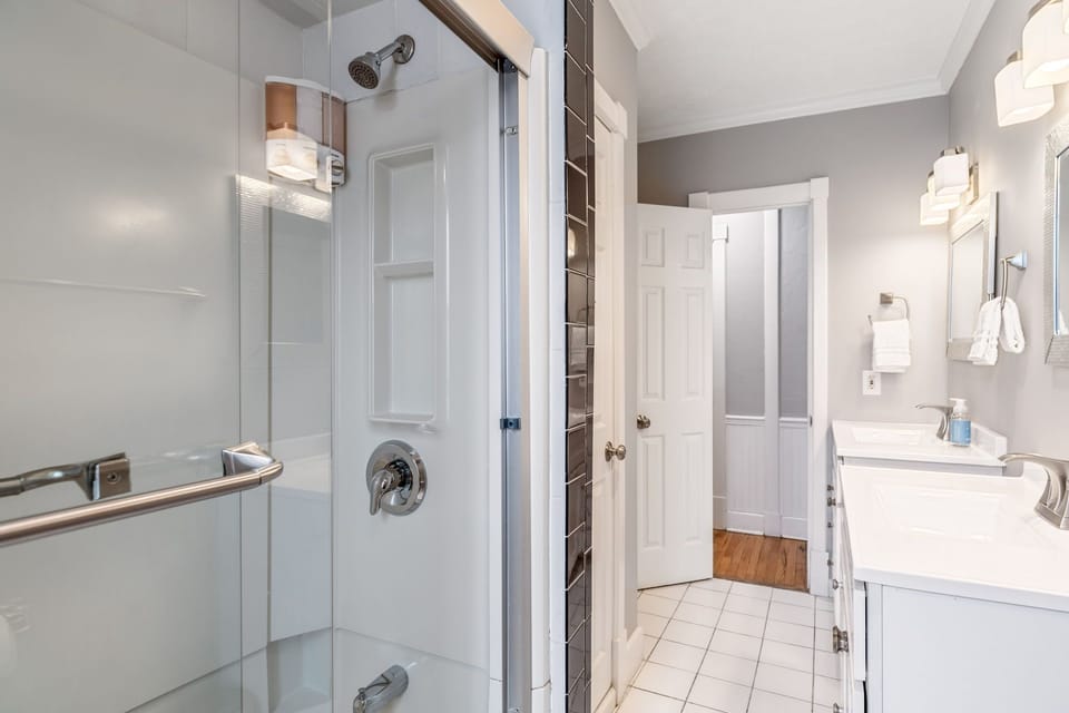 Bathroom with a glass-enclosed shower, two separate white sink vanities, tiled floor, wall mirrors, and bright vanity lights.
