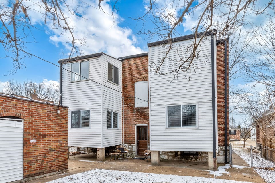 Rear view of the brick-and-white-sided multifamily building with a central exterior door, paved area, and snow-dusted yard.