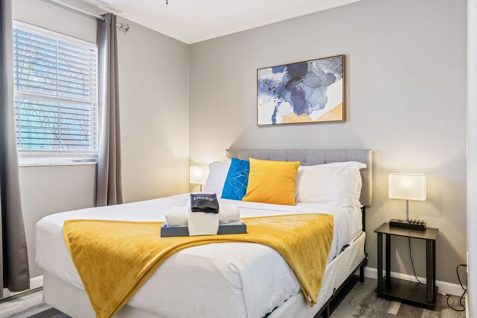 A gray upholstered bed features white bedding, a yellow throw, and blue and yellow accent pillows beneath abstract wall art.