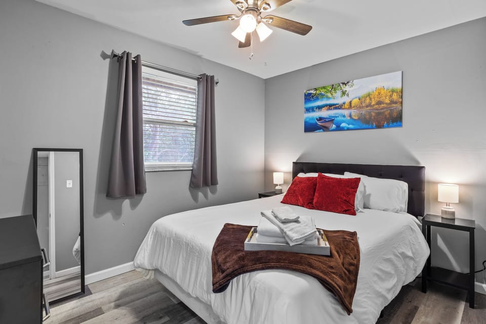 Daytime view of a gray bedroom with a made bed, red accent pillows, two nightstands, lamps, a window, dresser, and full-length mirror.