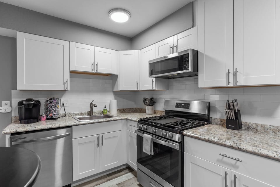 Close view of the kitchen with white cabinets, granite-look counters, a stainless steel gas range, microwave, dishwasher, sink, and coffee station.