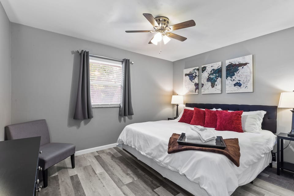 Daytime bedroom view with a king bed, white linens, red accent pillows, bedside lamps, world map artwork, a window, and a side chair.