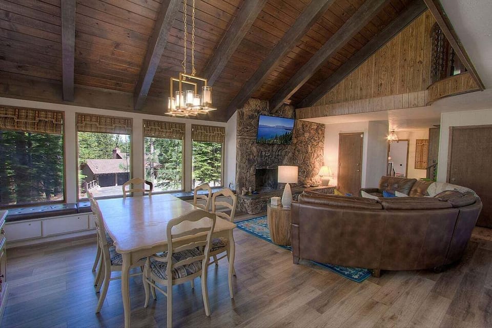 Dining area with table, chairs, chandelier, fireplace, TV, sectional sofa, large windows, window bench, and vaulted ceiling.