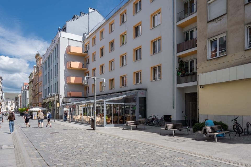 Another street view in a bustling city area, showcasing historic buildings, open pathways, and shops under a bright blue sky.
