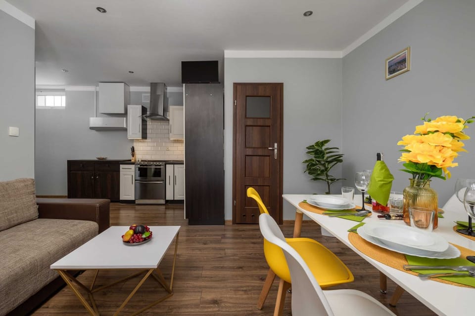 The living room features a cozy gray sofa accented with yellow pillows, bright yellow curtains, and a small dining table set for two. A vase with fresh flowers adds a decorative touch, creating a warm and inviting atmosphere.