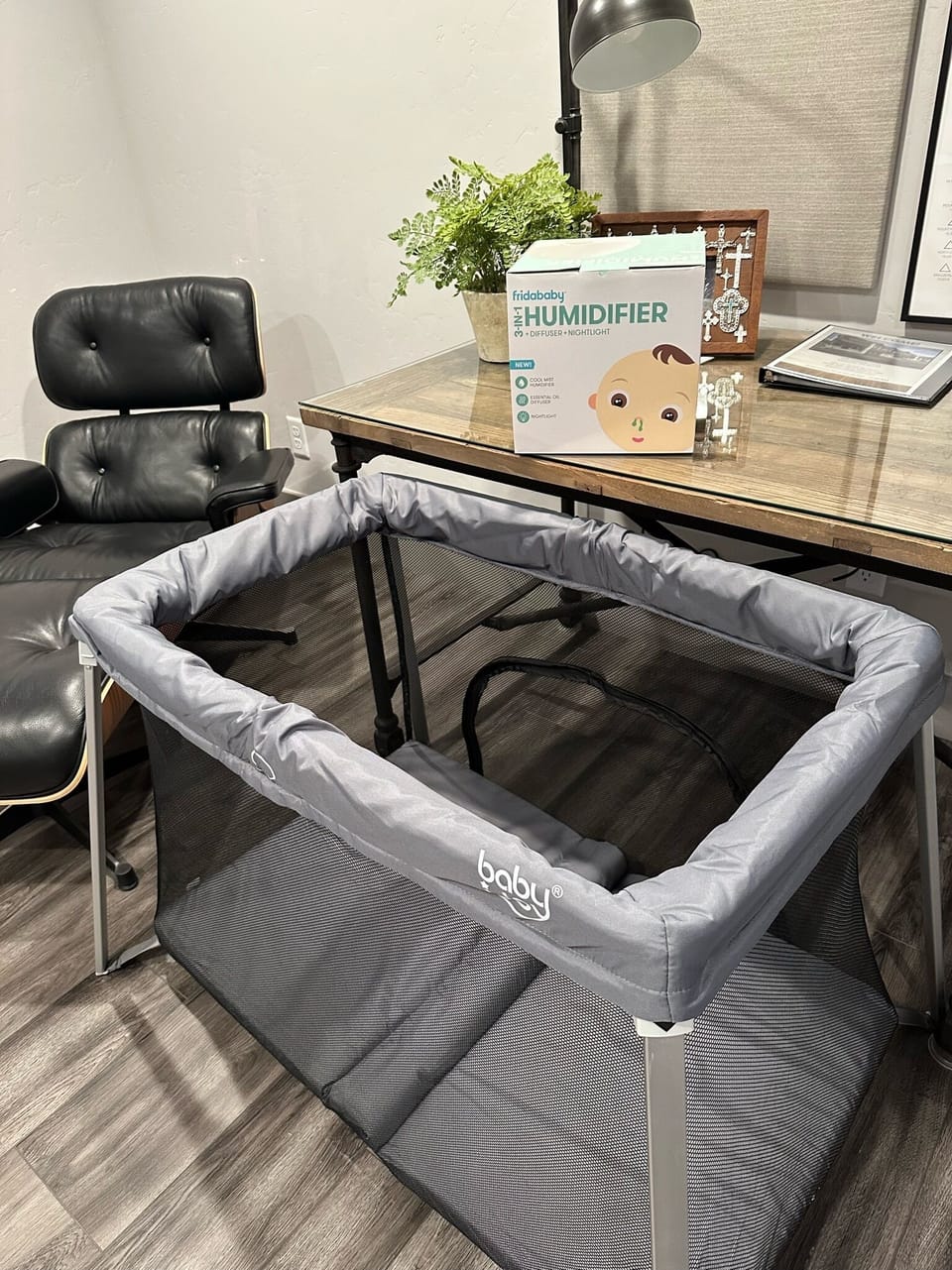 A gray infant playpen is set up on a wood floor next to a desk with a black chair. On the desk, there is a baby humidifier box and a potted plant.