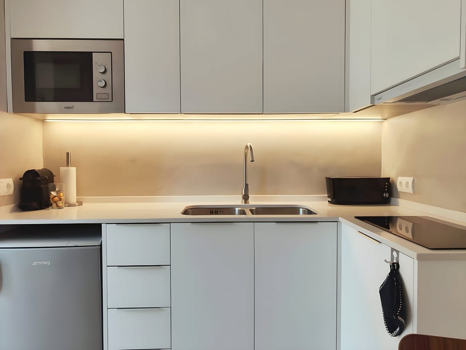 The modern, open, well-equipped kitchen features sleek white cabinets, a stainless steel sink, and a microwave. The kitchen is brightly lit with warm overhead lights, creating an inviting atmosphere for preparing meals.