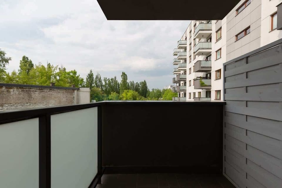A small outdoor balcony area with a dark railing and a view of green trees and nearby residential buildings.