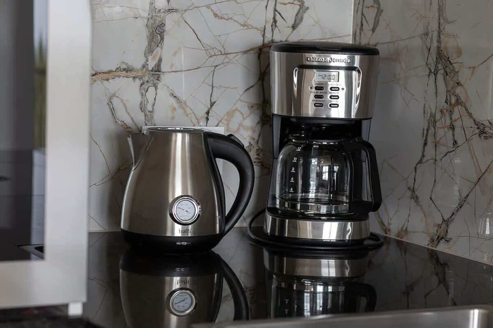 A coffee maker, electric kettle, and cups arranged on a marble countertop, perfect for a morning coffee setup.