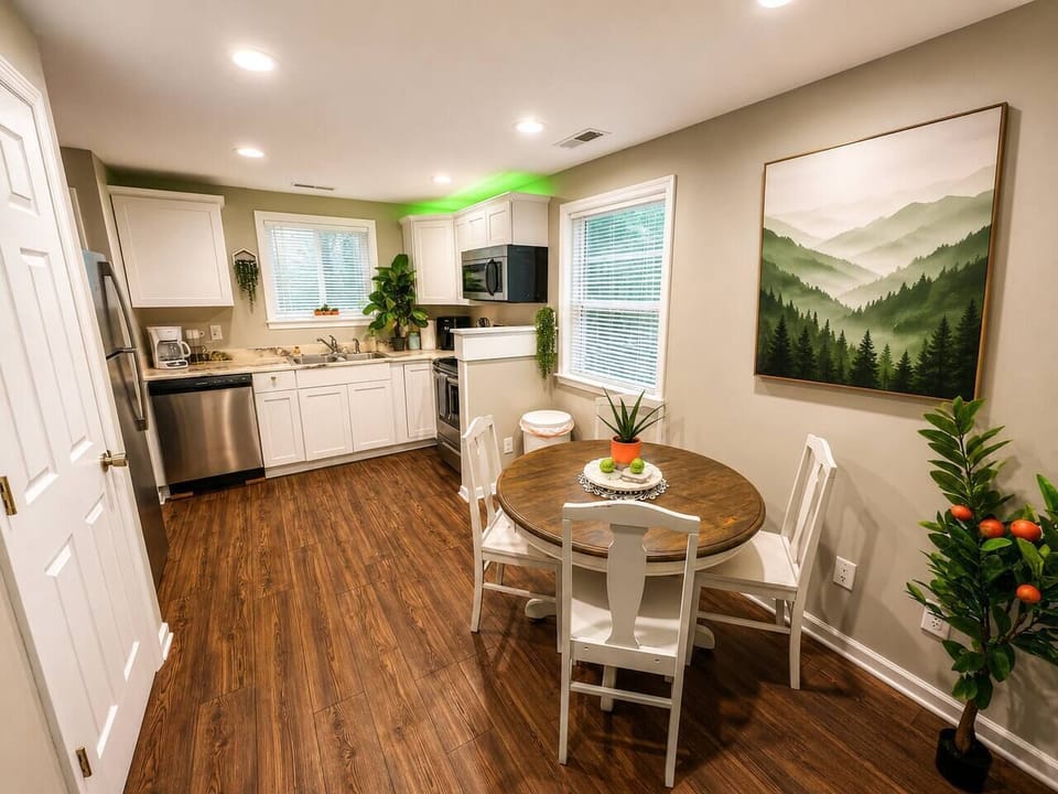 Compact kitchen and dining nook with white cabinets, stainless steel fridge & dishwasher, dark wood floors, and a round wood dining table with chairs. Cozy, functional setup for cooking, eating, or chilling over coffee.