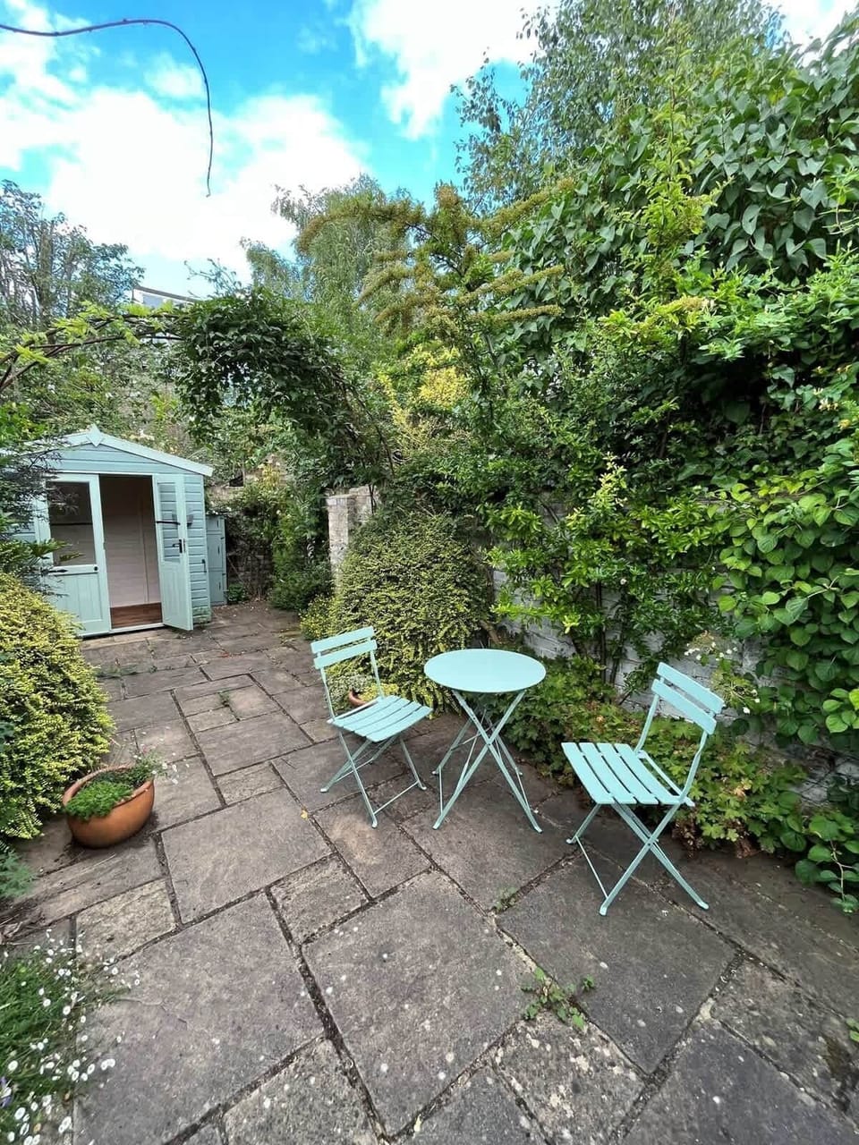 A charming, private stone courtyard surrounded by lush greenery, featuring a quiet bistro set and a quaint garden shed for a peaceful outdoor escape.