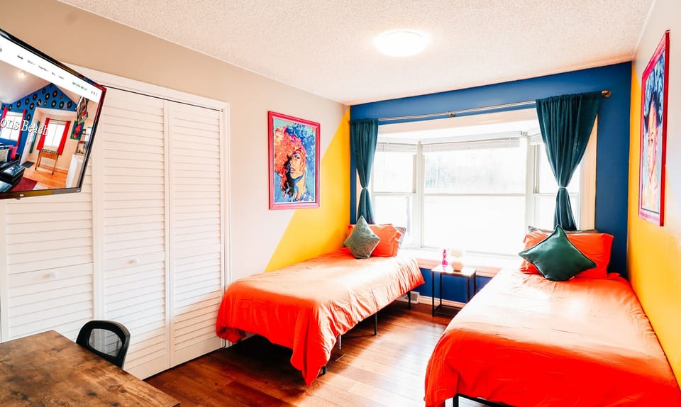 Twin Tranquility: Embrace Comfort in this Refreshing Bedroom with Twin Beds, Bathed in Vibrant Orange Hues and Complemented by Cool Bluish-Green Curtains. Enjoy the View and Entertainment with a Window and TV Screen 🛏️🪟📺