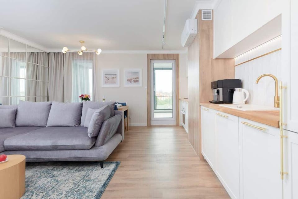 A living room with elegant decor, including a sectional sofa, framed artwork, and a large dining table. The space is brightened by natural light.