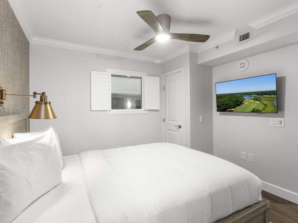 Drift off in your serene bedroom featuring crisp white linens, modern ceiling fan, and wall-mounted TV for cozy evening entertainment.