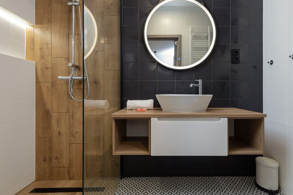 A bathroom with dark wall tiles, a round mirror, a wooden vanity, and a ceramic basin, exuding a chic and contemporary vibe.