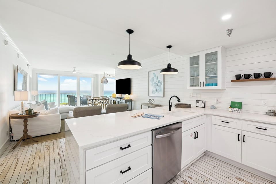 Your bright coastal kitchen flows seamlessly into the living space, where ocean views through floor-to-ceiling windows create the perfect backdrop for cooking.