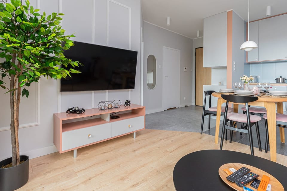 A contemporary living area with a flat-screen TV, a dining table, and chairs. A plant and shelves add elegance to the decor.