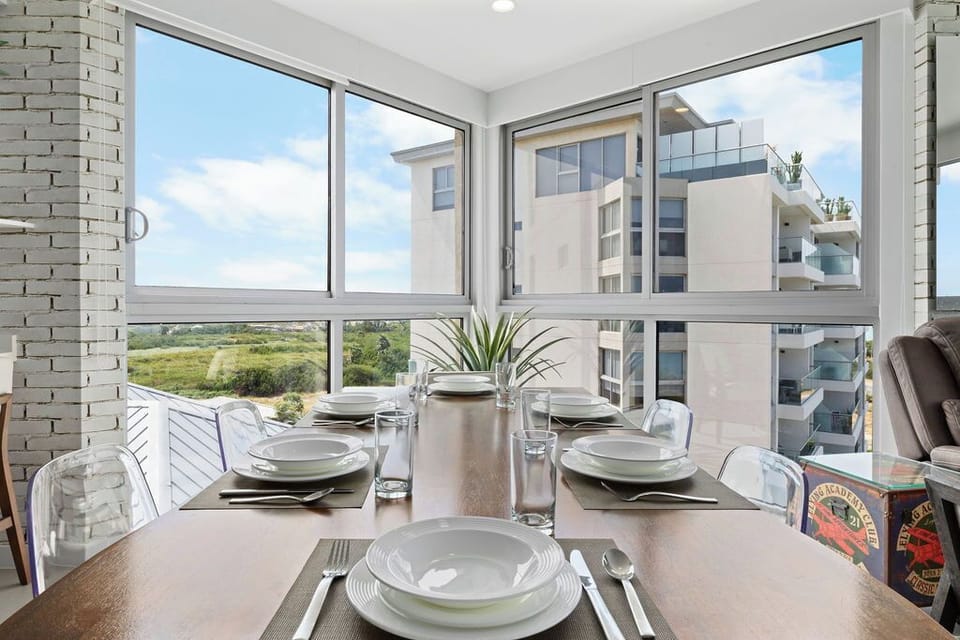 Savor meals at your elegantly set dining table with natural light streaming through expansive windows, framing views of greenery and contemporary architecture beyond.
