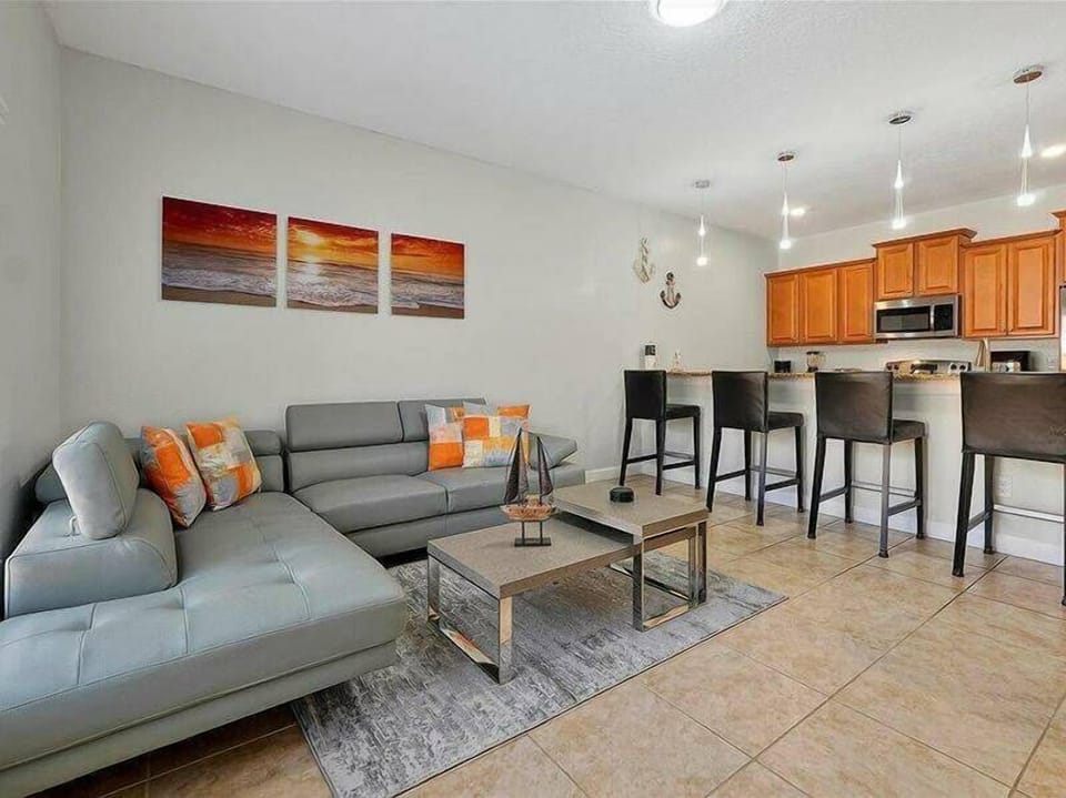 This inviting living area boasts a chic gray sectional with lively orange accents, fostering a cheerful ambiance. The sleek kitchen island is ideal for casual dining, complemented by three pendant lights that create a warm glow.