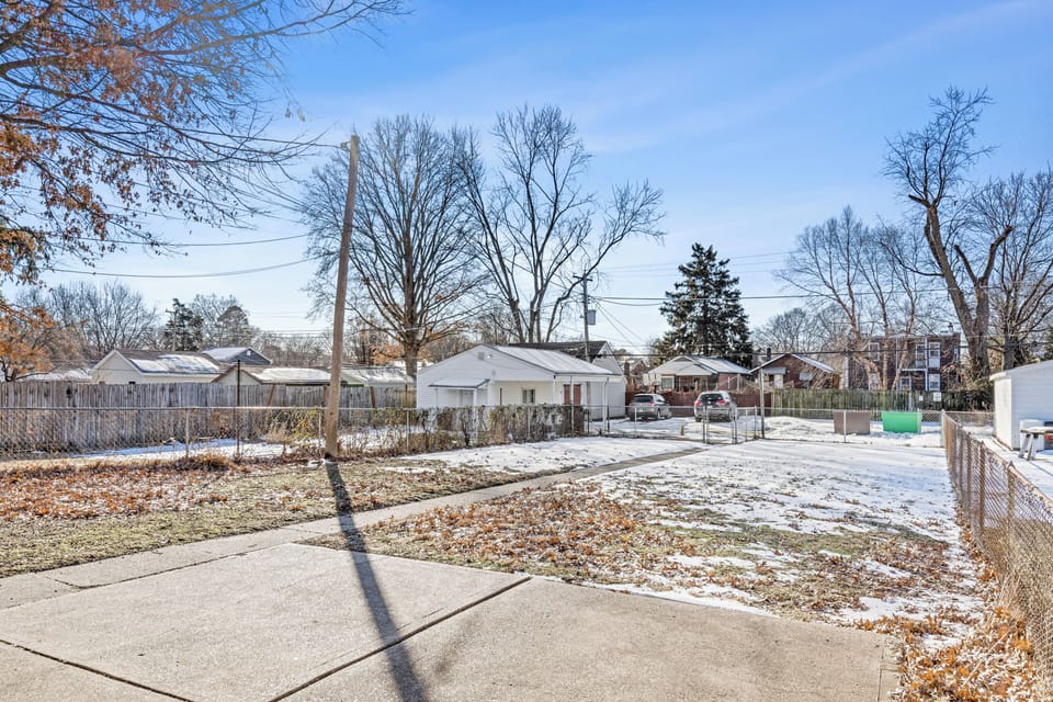 Wide winter view of the fenced backyard with concrete walkway, patchy snow, alley gate, garages, neighboring buildings, and utility lines.
