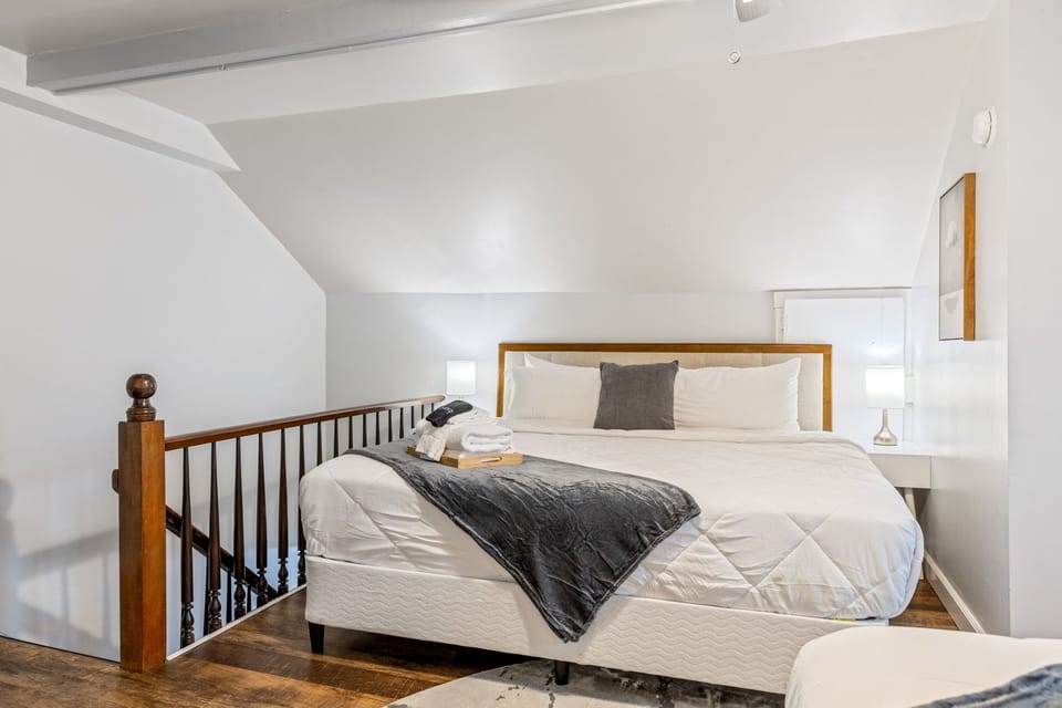 Loft bedroom with a large white bed, gray throw, bedside lamps, wood flooring, and an open railing beside the stairwell.