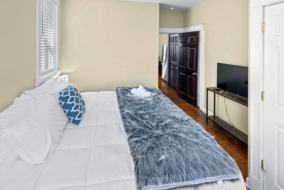 This reverse bedroom angle shows the white-dressed bed with gray throw, TV on a wood console, hardwood floors, windows, dark wood doors, and a view toward the kitchen.