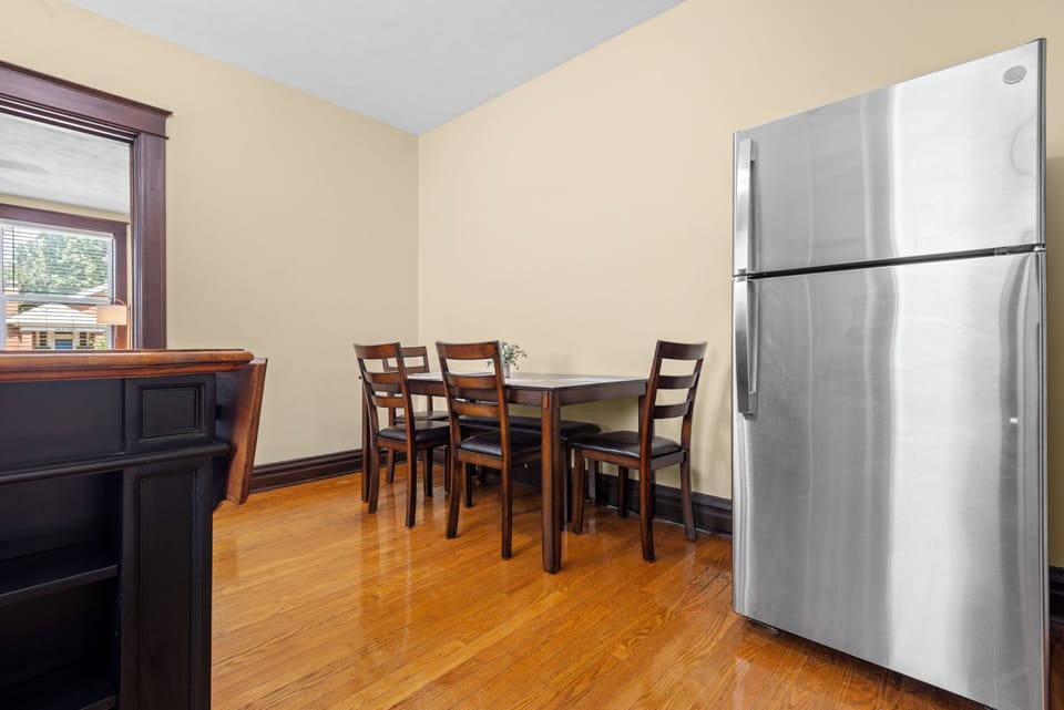 The dining area includes a four-place wood table beside a stainless refrigerator, hardwood floors, and the dark kitchen island at the edge of the room.