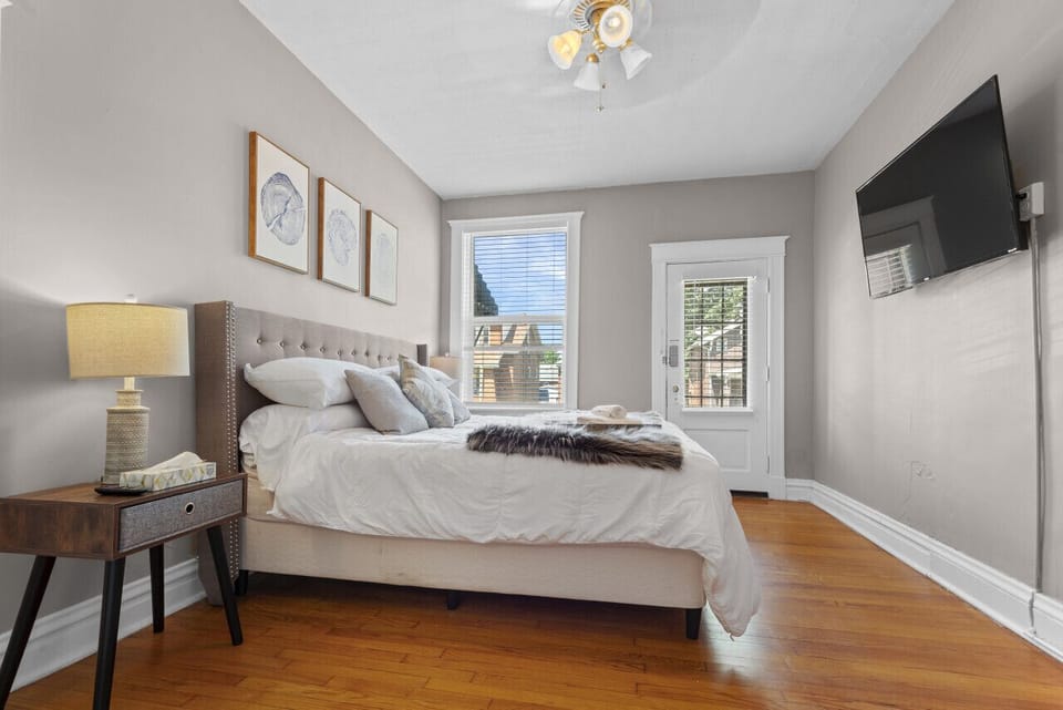 The bedroom features a white-dressed bed, gray tufted headboard, wall-mounted TV, nightstand, ceiling fan, window, and windowed door.