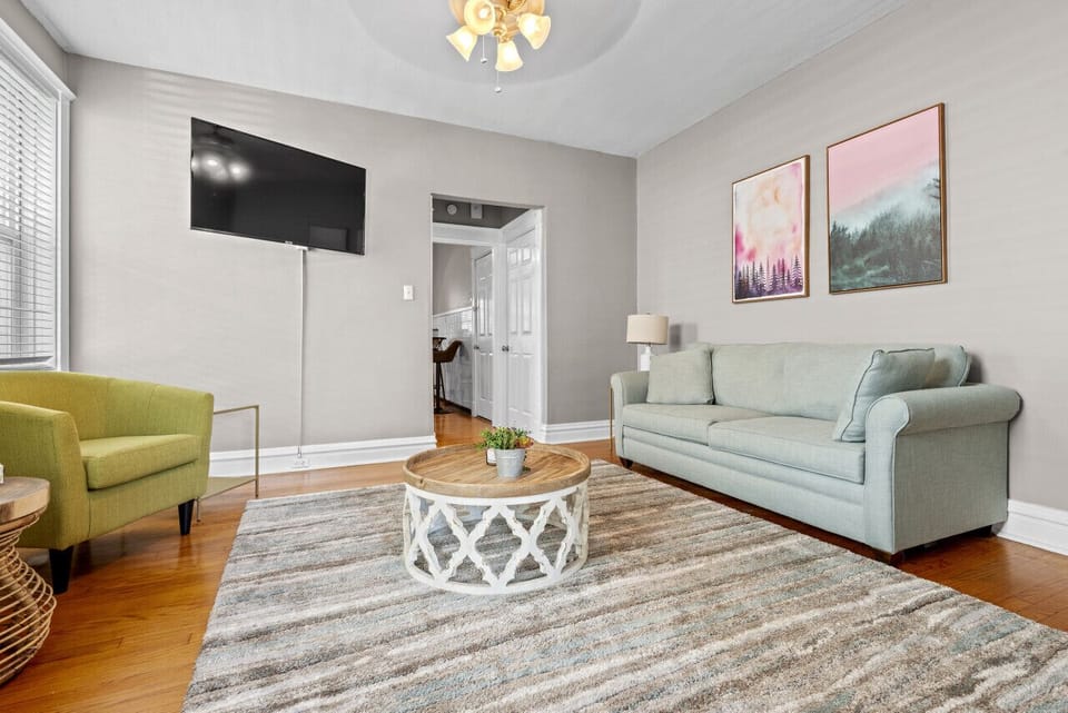 This reverse living-room view shows the light-gray sofa, green accent chair, wall-mounted TV, round coffee table, patterned rug, hardwood floors, and doorway toward the kitchen.