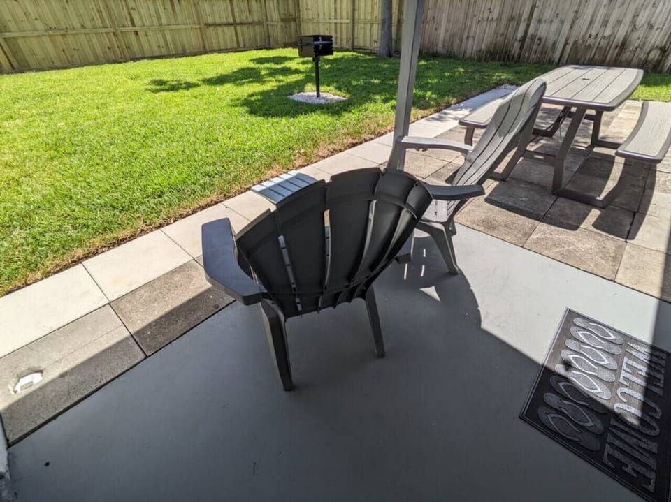 Another view of the lush green landscaped, private, fenced-in backyard with a large partially covered patio, outdoor dining area, Adirondack chairs, outdoor patio lighting, and grill.