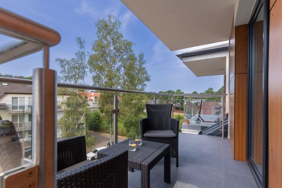 A balcony with a wooden railing and outdoor seating, overlooking greenery and trees.