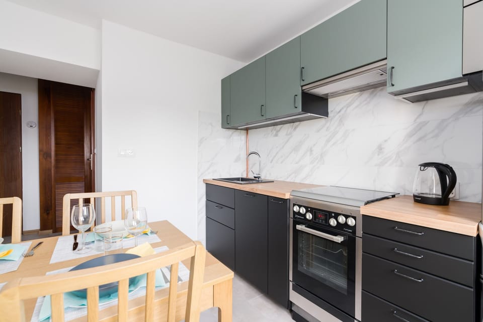 A modern kitchen with green cabinetry, marble countertops, and a dining table visible in the background.