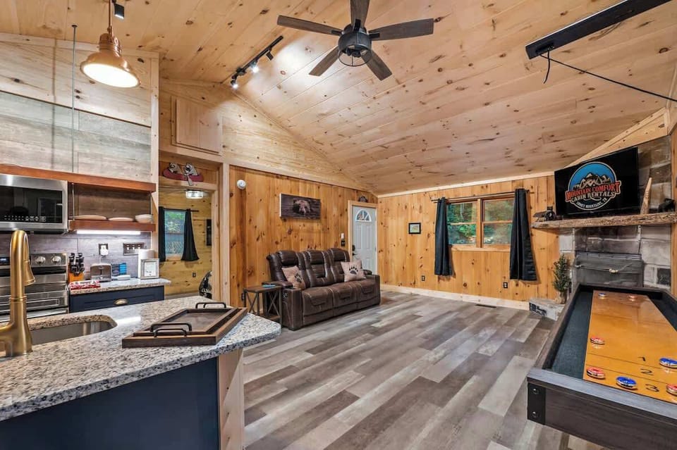 Spacious open-concept living area with cozy recliners, a stone fireplace, shuffleboard table, and warm wood finishes throughout.