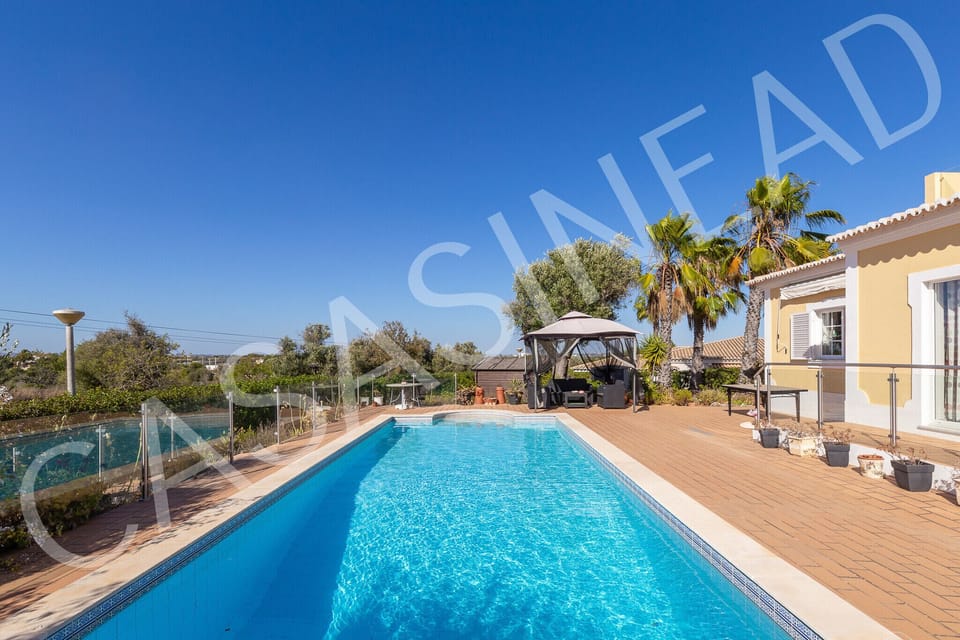 Crystal-clear swimming pool at Casa Sinead viewed from the wooden sun deck, with comfortable outdoor seating area, tropical palm trees, and the elegant villa facade visible in the background, perfect for relaxing holiday afternoons in the Algarve sunshine