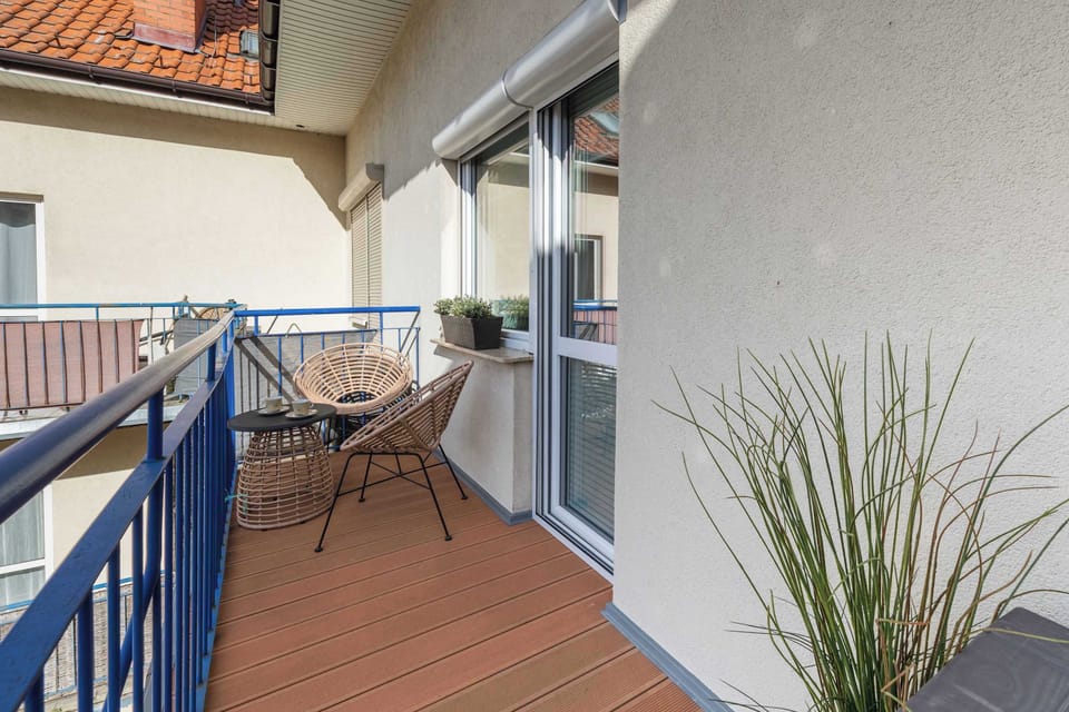 A small outdoor balcony with two chairs, a table, and a potted plant, overlooking the neighboring buildings.