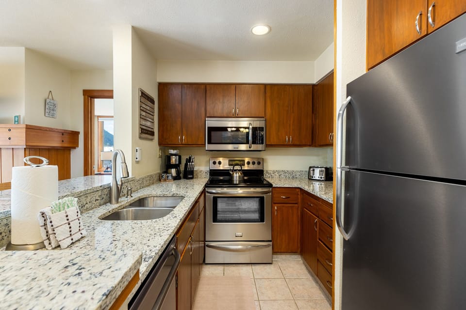 A modern kitchen with stainless steel appliances, granite countertops, a double sink, wooden cabinets, and a tile floor.