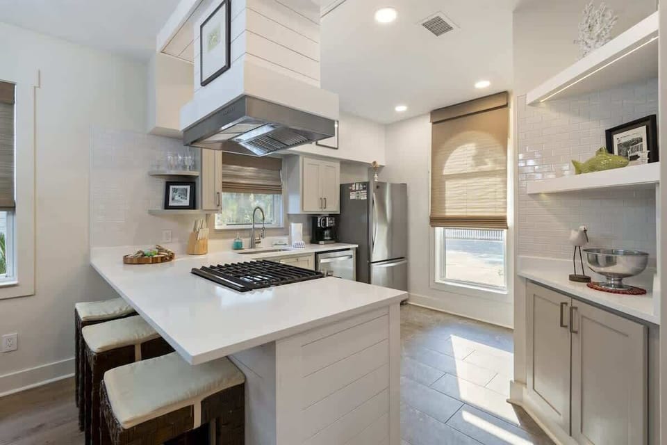 - Spacious kitchen island with bar seating creates a social hub for morning coffee - Sunlight pours in through windows, highlighting the crisp white finishes & wood accents - Open shelving & stylish range hood add modern charm & function to the space