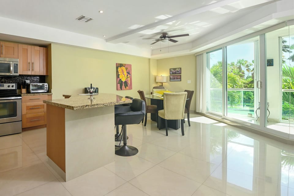 Step into your bright, open-plan living space where modern comfort meets tropical elegance. The sleek kitchen flows seamlessly into a stylish dining area, all bathed in natural light from floor-to-ceiling glass doors opening to lush greenery.