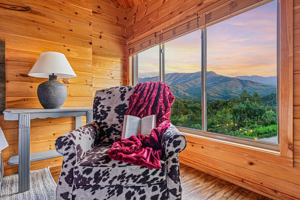 The upper level master bedroom frames an unobstructed panoramic view of the Smoky Mountains through large windows — wake up to this every morning. Soft morning light, the ridgeline stretching before you, and nowhere you need to be. This is what a mountain 