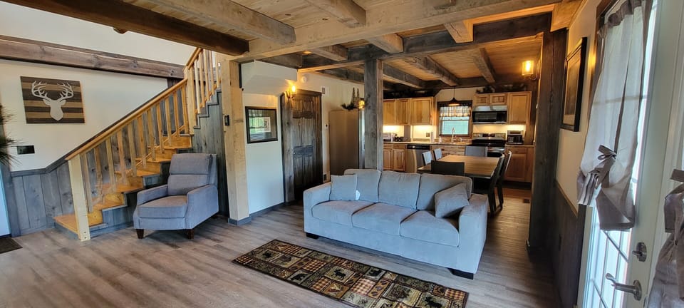 Spacious and cozy living room with doors to deck with couch and chair. Dining Area and Kitchen behind with modern amenities and setting