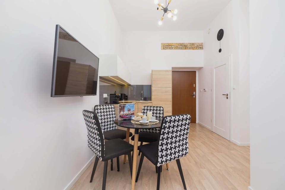 A dining area with a table and four chairs, complemented by a wall-mounted TV. The white walls and wooden floor create a clean and modern look.