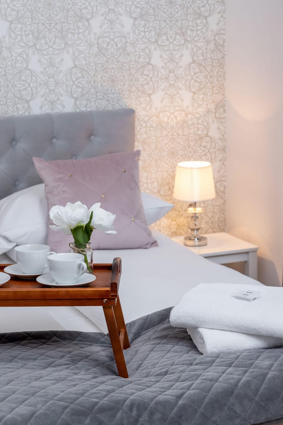 A cozy bedroom featuring a white bedspread and soft, diffused lighting. A single nightstand and minimal artwork maintain a serene setting.