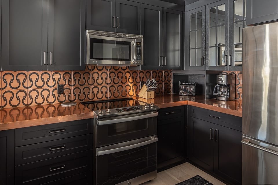 Step into your gourmet kitchen featuring sleek dark cabinetry, warm copper accents, and premium stainless steel appliances for memorable meal preparation.