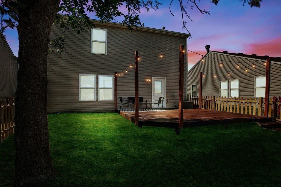 Fully fenced back yard with patio furniture, string lights and BBQ grill.