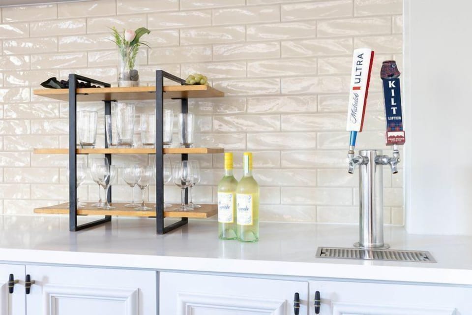 Beverage and wine display shelf with glassware, white wine bottles, and a stainless steel beer tap beside a tiled wall