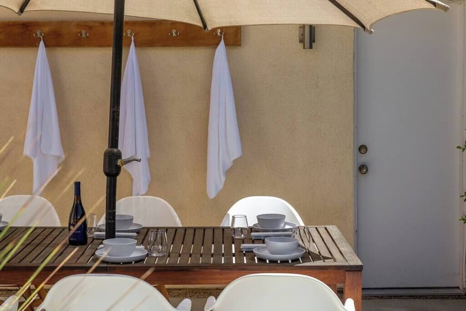 Shaded outdoor dining table set for six with patio umbrella, white towel hooks, and poolside access — alfresco meals made easy.