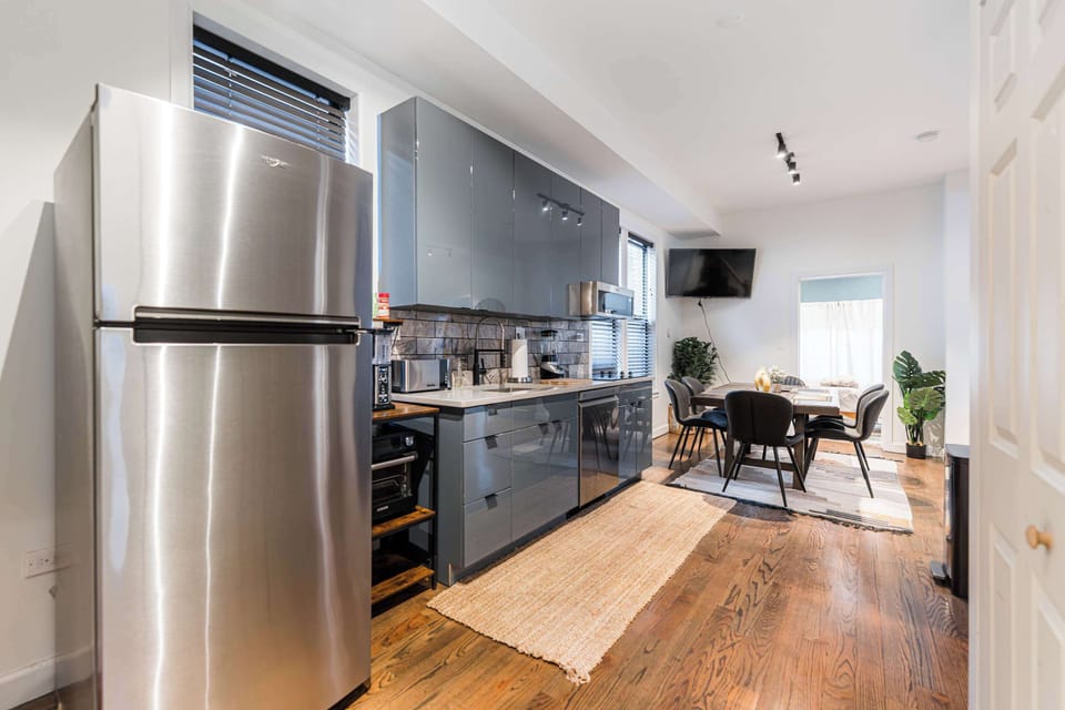 Modern kitchen and dining area with stainless steel fridge, sleek cabinetry, and warm wood floors. Fully equipped for cooking and entertaining, with a cozy dining table perfect for meals or working. A stylish, sunlit space designed for comfort