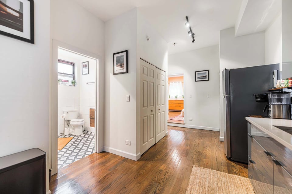 A bright, modern hallway with rich hardwood floors connects a sleek kitchen to a stylish bathroom featuring black-and-white patterned tile. Clean white walls, minimalist artwork, and recessed lighting create a warm, inviting, and open atmosphere.